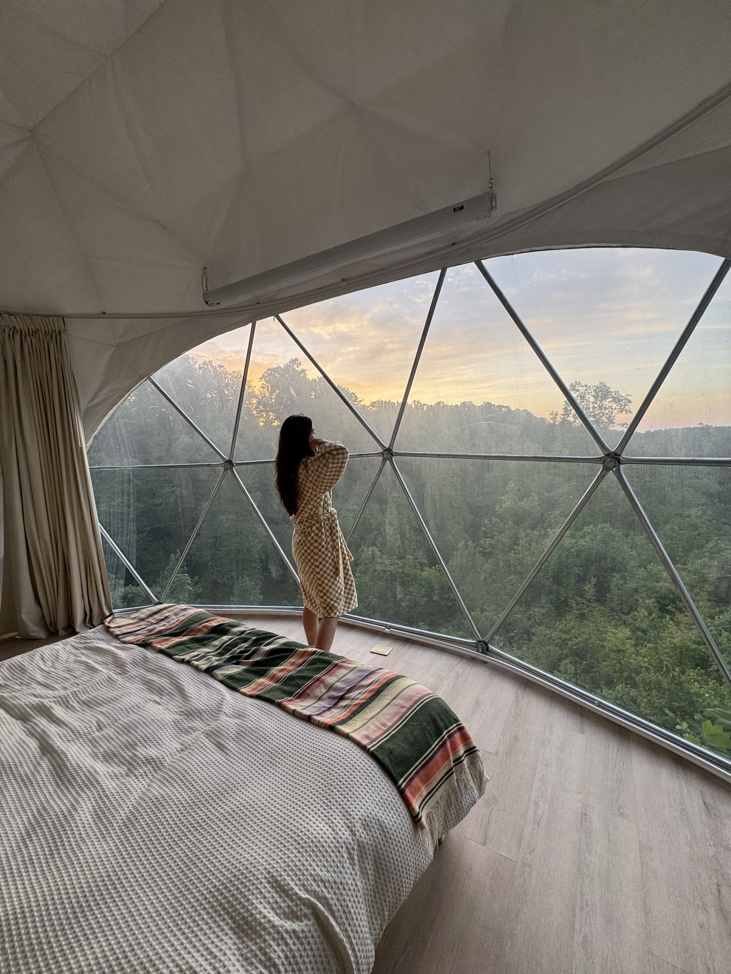 Guest in a robe looking out over the forest at sunrise through the curved geodesic window at Short Mountain Holiday