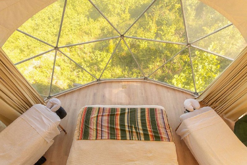 Couples massage setup inside the dome — two tables flanking the bed with the panoramic dome window looking out to the forest