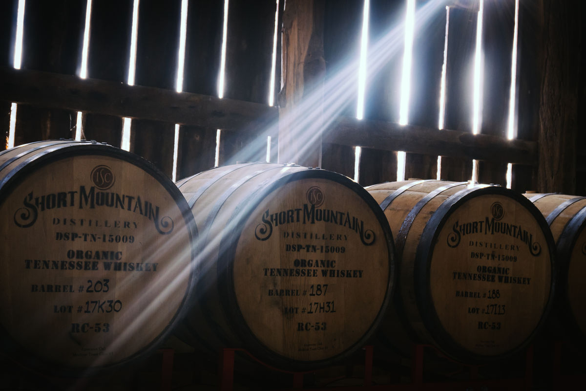 Short Mountain Distillery barrels in a wooden barn with sunlight streaming through the slats — Organic Tennessee Whiskey branded on each barrel head