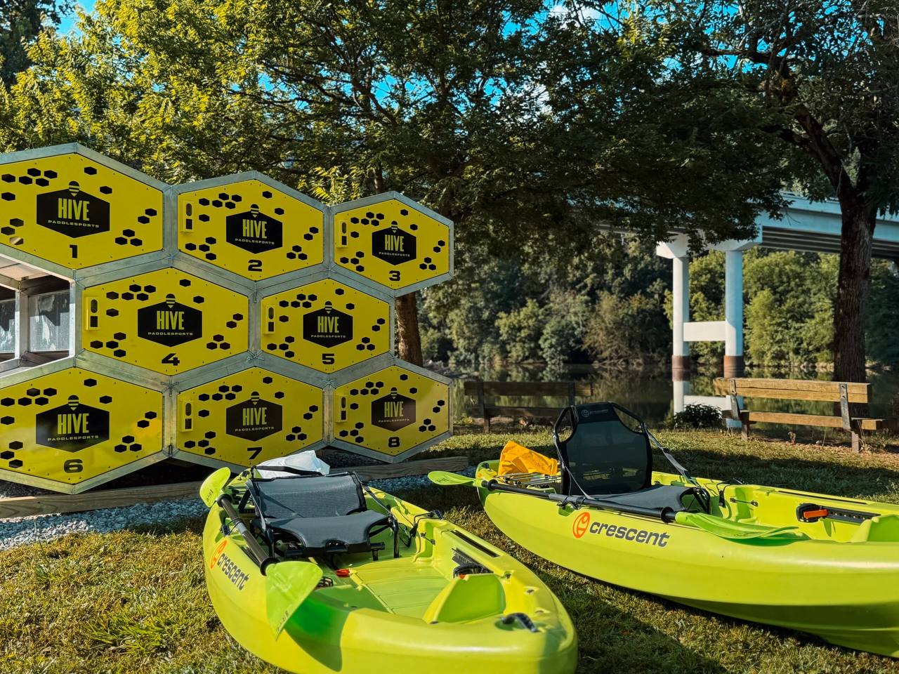 The Hive's numbered honeycomb kayak lockers beside two green kayaks on the riverbank at Riverfront Park in McMinnville, Tennessee